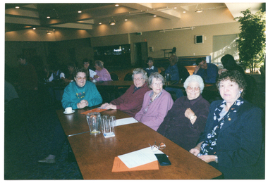 COL photo of numerous people seated at long rectangular tables in a large room. In the foreground, four women are seated at one of the tables. On top of the table, there are a jug and some glasses, a cup and saucer, some orange and some white papers, a pen, and some spectacles with their case.
