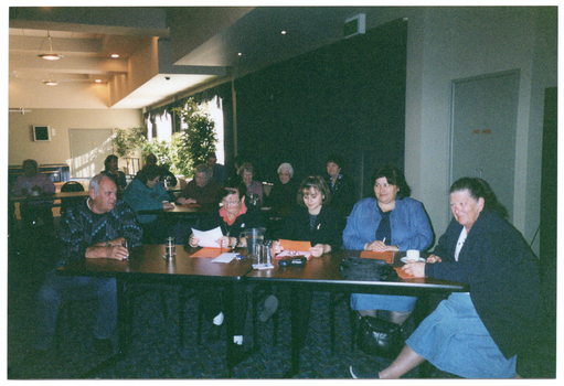 COL photo of numerous people seated at long rectangular tables in a large room. In the foreground, five people are seated at one of the tables. On top of the table, there are a jug and some glasses, a cup and saucer, some orange and some white papers, some spectacles with their cases, and a black shoulder bag. There is another black shoulder bag under the table.