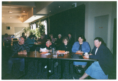 COL photo of numerous people seated at long rectangular tables in a large room. In the foreground, five people are seated at one of the tables. On top of the table, there are a jug and some glasses, a cup and saucer, some orange and some white papers, some spectacles with their cases, and a black shoulder bag. There is another black shoulder bag under the table.