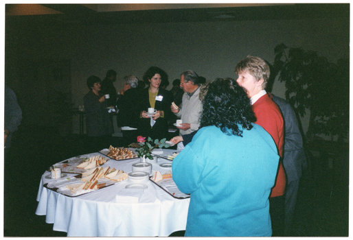 COL photo of a large group of people gathered around a room. In the foreground, several people are standing around a round table, on which there are platters of sandwiches, several stacks of small plates, paper napkins, a coffee cup and saucer, and a pink rose inside a small vase. In the background, there are people chatting to one another while holding cups and saucers.