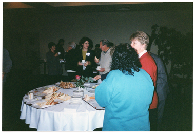 COL photo of a large group of people gathered around a room. In the foreground, several people are standing around a round table, on which there are platters of sandwiches, several stacks of small plates, paper napkins, a coffee cup and saucer, and a pink rose inside a small vase. In the background, there are people chatting to one another while holding cups and saucers.
