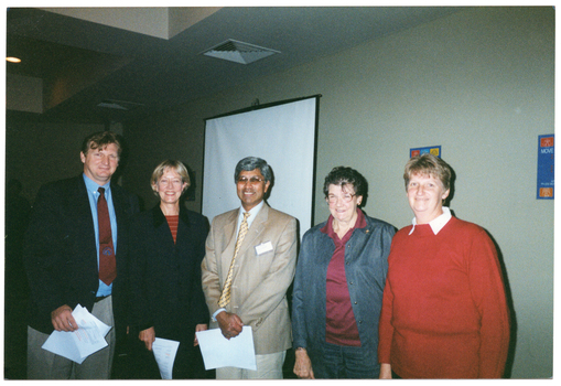 COL photo of five people, standing next to one another in a row. Three of them are holding some papers in their hands. In the background, there is a slide projector screen on a stand and some posters on the wall.