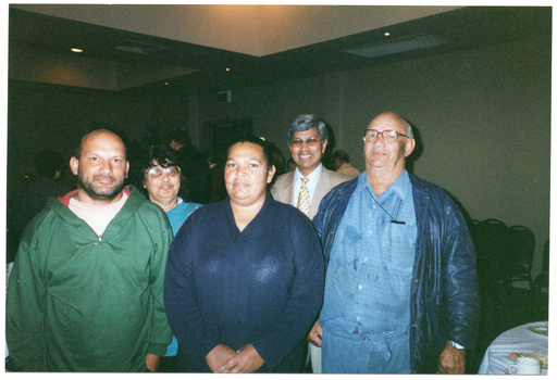 COL photo of a small group of people standing in a crowded room. They are facing the camera. Numerous other people, tables, and chairs are evident in the background.