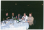 COL photo of a small group of people sitting at a round dinner table. One woman is holding a video cassette in a large plastic case. On the table, there are some small plates of food (such as cheese and fruit, cakes, and bread rolls), a bottle of wine, a water jug, cups and saucers, wine, beer, and water glasses, a pair of salt and pepper shakers, a bottle and a can of soft drink, some pens, and some large booklets and notepads. In the centre, there is a small white vase of flowers. The background is dark, and there are a few downlights hanging from the ceiling.