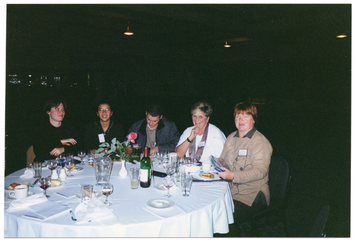 COL photo of a small group of people sitting at a round dinner table. One woman is holding a video cassette in a large plastic case. On the table, there are some small plates of food (such as cheese and fruit, cakes, and bread rolls), a bottle of wine, a water jug, cups and saucers, wine, beer, and water glasses, a pair of salt and pepper shakers, a bottle and a can of soft drink, some pens, and some large booklets and notepads. In the centre, there is a small white vase of flowers. The background is dark, and there are a few downlights hanging from the ceiling.