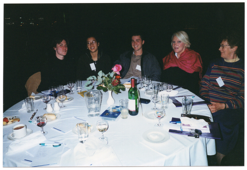 COL photo of a small group of people sitting at a round dinner table. In the centre of the table, there is a small white vase of flowers and a video cassette in a large plastic case. These are surrounded by some small plates of food (such as cheese and fruit, cakes, and bread rolls), a bottle of wine, a water jug, cups and saucers, wine, beer, and water glasses, a pair of salt and pepper shakers, some large booklets and notepads, and some pens. The background is dark.