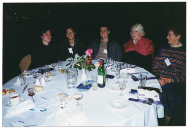 COL photo of a small group of people sitting at a round dinner table. In the centre of the table, there is a small white vase of flowers and a video cassette in a large plastic case. These are surrounded by some small plates of food (such as cheese and fruit, cakes, and bread rolls), a bottle of wine, a water jug, cups and saucers, wine, beer, and water glasses, a pair of salt and pepper shakers, some large booklets and notepads, and some pens. The background is dark.