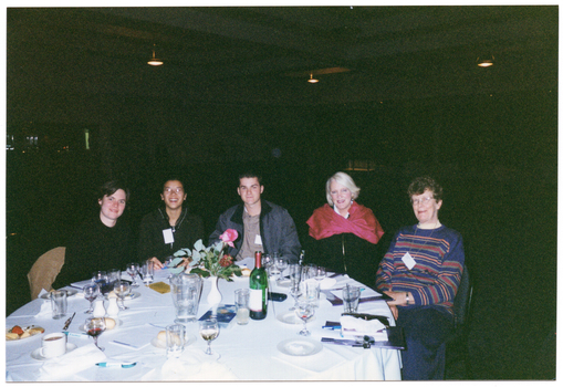 COL photo of a small group of people sitting at a round dinner table. On the table, there are some small plates of food (such as cheese and fruit, cakes, and bread rolls), a bottle of wine, a water jug, cups and saucers, wine, beer, and water glasses, a pair of salt and pepper shakers, a video cassette in its plastic case, some large booklets and notepads, and some pens. In the centre, there is a small white vase of flowers. The background is dark, and there are a few downlights hanging from the ceiling.