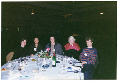 COL photo of a small group of people sitting at a round dinner table. On the table, there are some small plates of food (such as cheese and fruit, cakes, and bread rolls), a bottle of wine, a water jug, cups and saucers, wine, beer, and water glasses, a pair of salt and pepper shakers, a video cassette in its plastic case, some large booklets and notepads, and some pens. In the centre, there is a small white vase of flowers. The background is dark, and there are a few downlights hanging from the ceiling.