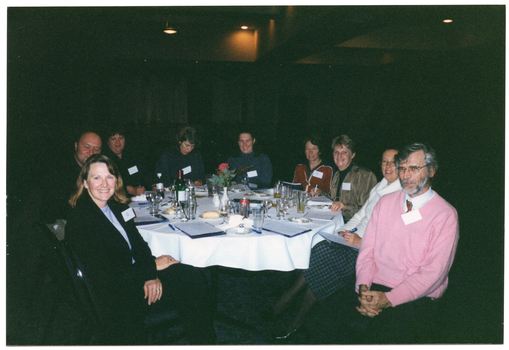 COL photo of a small group of people sitting at a round dinner table. On the table, there are some small plates of food (such as cheese and fruit, cakes, and bread rolls), a bottle of wine, a water jug, cups and saucers, wine, beer, and water glasses, a pair of salt and pepper shakers, a video cassette in its plastic case, some large booklets and notepads, and some pens. In the centre, there is a small white vase of flowers. The background is dark, and there are a few downlights hanging from the ceiling.