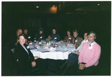 COL photo of a small group of people sitting at a round dinner table. On the table, there are some small plates of food (such as cheese and fruit, cakes, and bread rolls), a bottle of wine, a water jug, cups and saucers, wine, beer, and water glasses, a pair of salt and pepper shakers, a video cassette in its plastic case, some large booklets and notepads, and some pens. In the centre, there is a small white vase of flowers. The background is dark, and there are a few downlights hanging from the ceiling.