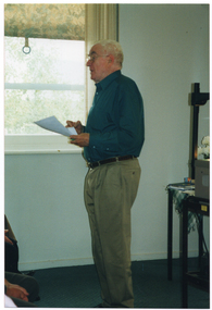 COL side view photo of a man standing near a window indoors. He is holding a piece of paper in one hand. Behind him, there is a small table (partially obscured) with some miscellaneous items on top. To the left of the frame, there are several people sitting on chairs (partially obscured), facing the man.
