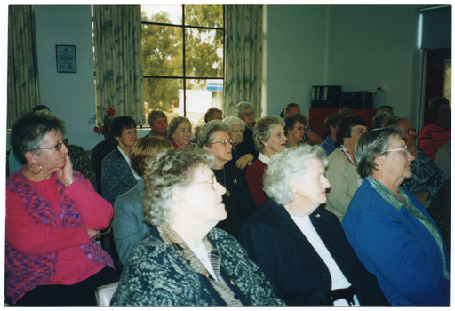 COL photo of a large group of people sitting on rows of chairs in a large room. They are looking forward, off camera. There is a small cupboard in the corner of the room, with some large folders and a portable stereo player on top. Behind the group, there are two curtained windows, through one of which several trees and a van with an open side door are evident outside.