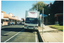 COL photo of a large van parked in a street parking space in a shopping strip. The text, "Healthy Bones Bus", has been painted above the front cabin. On the side of the van, there is a large blue awning and a wide door that is in the open position. There is also a set of stairs with a metal ballustrade coming out from the side of the van. Various commercial buildings are evident along the opposite side of the street.