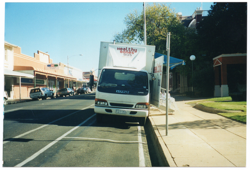 COL photo of a large van parked in a street parking space in a shopping strip. The text, "Healthy Bones Bus", has been painted above the front cabin. On the side of the van, there is a large blue awning and a wide door that is in the open position. There is also a set of stairs with a metal ballustrade coming out from the side of the van. Various commercial buildings are evident along the opposite side of the street.