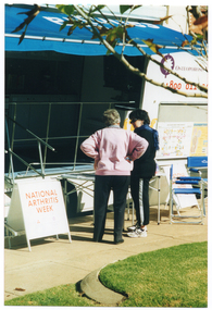 COL photo of two women, standing, facing a large van which has been parked adjacent to a paved area. Above them, there is a large blue awning jutting out from the van, at roof level. Between the van and the women, there is a rectangular table. The side of the van is completely open with a metal ballustrade in place. There is a large side door on the right that is in an open position. It contains the text, "OSTEOPOROSIS..." (partially obscured), a telephone number, "1800 011..." (partially obscured), and a display board affixed to the door below the text. There are two white A-frame sandwich boards on either side, on the paved area in front the vans. The sandwhich boards contain the text, "NATIONAL ARTHRITIS WEEK", painted in red. Two small logos are evident beneath the main text. Behind the sandwich board on the left, there is a set of metal steps with a metal ballustrade, leading into the van. In front of the sandwich board on the right, there are two blue deckchairs. In the foreground, the branches of a tree evident at the top of the photo image and, at the bottom of the image, there is a grassy area beyond the paving.