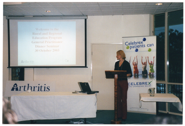 COL photo of a woman standing behind a wooden lectern. Beside her, there is a large slide projector screen hanging from the wall, and a large rectangular table beneath it. The woman is looking behind towards the screen. The table is covered with a white tablecloth and has a laptop computer at one end. A slide being projected onto the screen contains the text, "Welcome to the Rural and Regional Education Program General Practitioner Dinner Seminar 30 October 2003". In the two bottom corners of the slide, there is a logo for Arthritis Victoria, and one for Osteoporosis Victoria. Propped up behind the table, there is a large "Arthritis Victoria" sign. On the other side of the woman, there is a small table, also covered with a white tablecloth. It has a glass jug of water on top. Behind the woman, there is a large, freestanding, vertical banner containing the text, "Celebrex patients can", with two green and two blue circles or balls around the text. Beneath the text, there is a picture of four people standing thigh deep in the sea (one partially obscured), with their arms raised above their heads. Below the picture, there is the text, "CELEBREX LIFE CHANGING RELIEF". Behind the banner, there is a closed door to the left and, to the right, there are some glass floor to ceiling windows. Through the windows, a car park and some large trees in the distance are evident at dusk.