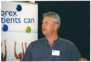 COL close up photo of a man in three quarter profile. He is wearing a name tag on his shirt. Behind him, there is a vertical banner containing the text, "Celebrex patients can" (partially obscured) with one green and two blue circles or balls around the text. Beneath the text, there is a picture of a few people standing thigh deep in the sea (partially obscured), with their arms raised above their heads.