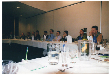 COL wide frame photo of a group of people sitting at several long rectangular tables which have been joined together in a u-shape. The tables are all covered with white tablecloths. On top of the tables, in the foreground, there is a glass jug half filled with water, an empty beer glass and an empty wine glass, a bottle of wine in a clear plastic wine cooler, and some printed paper on top of a plate, with some pens placed on top. Similar items are on the other tables in the background.