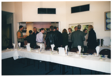 COL photo of a group of people standing close together in a room, drinking and chatting. They are standing between a bar and some long rectangular tables covered with white tablecloths. There are various items on the tables, such as wine glasses, cutlery, some printed publications, some pens inside a holder, salt and pepper shakers, and two table-top display stands.
