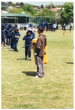 COL photo of a man and a group of school children on a grassy sports field. The man is holding a large yellow padded shield. He is facing the school children, who are queueing up while one student begins to towards the man. In the backgroud, there is one person walking away from the group. In the distance, several students are gathered in groups around the field. There is a small building on the school grounds and, beyond that, some houses and trees.