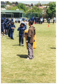 COL photo of a man and a group of school children on a grassy sports field. The man is holding a large yellow padded shield. He is facing the school children, who are queueing up while one student begins to towards the man. In the backgroud, there is one person walking away from the group. In the distance, several students are gathered in groups around the field. There is a small building on the school grounds and, beyond that, some houses and trees.