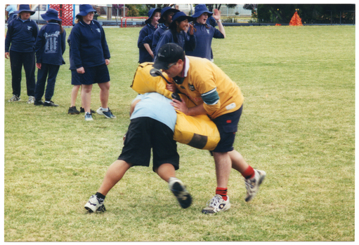 COL photo of a school student tackling a man who is holding a yellow padded shield. They are on a grassy sports field. The student is bending forward, and the man has the shield across the student's back. There are several other students standing in a queue nearby. Some of them are looking off camera and laughing.