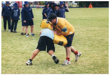 COL photo of a school student tackling a man who is holding a yellow padded shield. They are on a grassy sports field. The student is bending forward, and the man has the shield across the student's back. There are several other students standing in a queue nearby. Some of them are looking off camera and laughing.