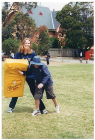 COL photo of an adult instructor and a school student practising tackling, using a large yellow padded shield. They each have one leg crossed over the other's leg, and the student is pushing one arm into the shield. The instructor is wearing a blue collared t-shirt with the Arthritis Victoria/Osteoporosis Victoria logos in yellow. The shield has the "Bundaberg Rum" branding and logo on it. In the background, there are several students across a gravel or concrete area beyond sporting field, including one student who is holding a basketball. Behind them, there is a large ornate brick building with a steep roof.