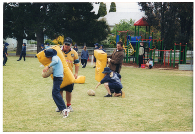 COL photo of two adult instructors and some school children on a grassy sports field. The instructors are each holding a padded yellow shield. One of the students is lunging into the shield while holding a football. The other student is reaching for a football on the ground. In the distance, several school children are standing on the field or in front of a playground area with woodchips on the ground. In the background, there are some buildings and trees.