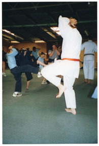 COL photo of a group of school students and two martial arts instructors in a large carpeted room. The students are wearing their school uniforms and are practising  some kicks while they hold their hands on their heads. The two adult instructors are wearing white martial arts uniforms, one with an orange belt and one with a black belt. One of the instructors is demonstrating the kicking practice exercise to the students.
