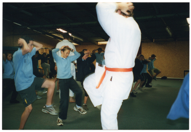COL photo of a group of school students and two martial arts instructors in a large carpeted room. The students are wearing their school uniforms and the instructors are wearing a white martial arts uniforms, one with an orange belt and one with a black belt. One instructor is demonstrating a kicking technique while the other instructor walks around the class, observing the students. They are kicking with one leg while holding their hands on their heads.