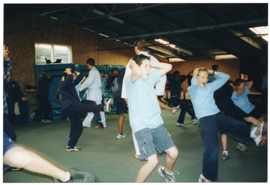 COL photo of a group of school students and two martial arts instructors in a large carpeted room. The students are wearing their school uniforms and the instructors are wearing a white martial arts uniforms, one with a white belt. One instructors is walking around the room while the other is standing in one place. The students are practising kicking with one leg while holding their hands on their heads. In the background, there are some large padded gym mats leaning against the wall amongst some other equipment.