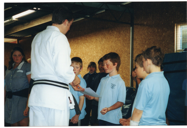 COL photo of a small group of students and a martial arts instrucstor standing inside a room. The instructor is handing out a piece of paper to a student, who is smiling as he accepts it while the other students look on.