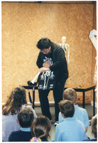 COL photo of a woman standing in front of a small group of school children, who are sitting on the floor. The woman is wearing a black apron with a white skeleton drawing on it. In the bottom left corner of the apron, there is the text, "BONES & JOINTS". The woman is bending forward, looking at the apron as she positions a plastic bone above the skeleton drawing. There is a plastic skeleton on a stand perched on a table behind her, and another larger skeleton on a stand beside her (partially obscured).