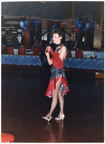 COL photo of a woman standing on the dance floor of a large hall. She is dressed in a red and grey dance costume, and is speaking into a cordless hand-held microphone. In the background, there are several tables and chairs set up in a row. A few people are standing to one side between some tables, while some others are sitting at some tables. There are red, white, and blue balloons floating above the tables, and some LED lighting strips decorating the columns and other architectural features.