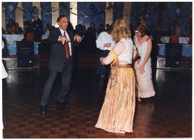 COL photo of a small group of people dancing on a dance floor. Two of the people are dressed in traditional dance costumes, one person is wearing a suit and tie, and another person is wearing a white shirt and checked skirt. Around the edge of the dance floor, there are two "Move It or Lose It" banners affixed to a low partition wall separating the dining area from the dancefloor. In the background, there are several people sitting at tables in the dining area, some of whom are watching the dancers. There are red, white, and blue balloons floating above the tables and streamers decorating the walls.