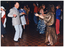 COL photo of several people dancing on a crowded dance floor. Some people are dressed in traditional dance costumes while others are dressed in contemporary clothes. In the foreground, there is a man wearing a light grey suit and tie dancing with a woman wearing a long metallic gold dress with a black and gold scarf around her hips.