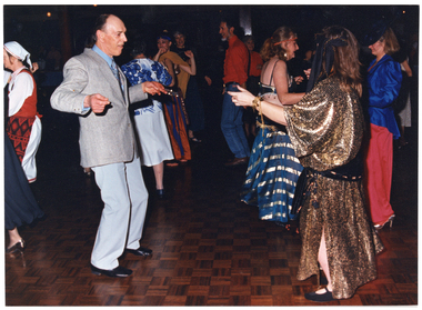 COL photo of several people dancing on a crowded dance floor. Some people are dressed in traditional dance costumes while others are dressed in contemporary clothes. In the foreground, there is a man wearing a light grey suit and tie dancing with a woman wearing a long metallic gold dress with a black and gold scarf around her hips.