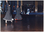 COL photo of two women dancing on a dance floor in a large hall. They are both dressed in long-sleeved polka dot dresses, with a frill at the hem. In the background, to the left of the frame, there are a few people sitting at tables, and a waitperson carrying a tray. To the centre right of the frame, there is a closed double doorway with a green "EXIT" sign above it. The hall has been decorated with balloons, streamers, and LED light strips.