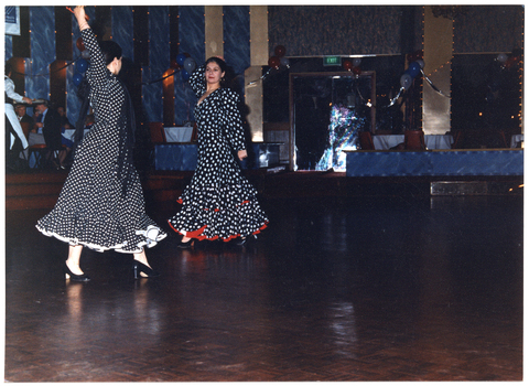 COL photo of two women dancing on a dance floor in a large hall. They are both dressed in long-sleeved polka dot dresses, with a frill at the hem. In the background, to the left of the frame, there are a few people sitting at tables, and a waitperson carrying a tray. To the centre right of the frame, there is a closed double doorway with a green "EXIT" sign above it. The hall has been decorated with balloons, streamers, and LED light strips.