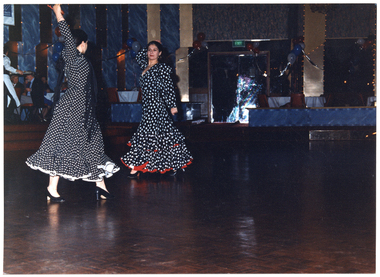 COL photo of two women dancing on a dance floor in a large hall. They are both dressed in long-sleeved polka dot dresses, with a frill at the hem. In the background, to the left of the frame, there are a few people sitting at tables, and a waitperson carrying a tray. To the centre right of the frame, there is a closed double doorway with a green "EXIT" sign above it. The hall has been decorated with balloons, streamers, and LED light strips.