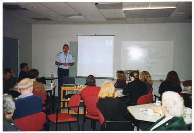 COL photo of a police officer speaking to a group of people inside a large room. Behind him, there is a freestanding slide projector screen on a stand. In front of him, there is a small table with a laptop and slide projector on top. Beside the screen, there is a large whiteboard affixed to the wall. Small groups of people are seated around small hexagonal tables, on which there are cups and saucers and plates of food.