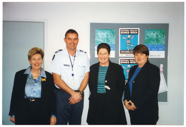 COL photo of four people standing next to one another in a row. One of them is a police officer in uniform. Behind them, there is a large display board affixed to the wall. There are some posters on the display board, including two identical "Does arthritis limit your life?" posters (in blue, black and white), and two identical "National Healthy Bones Week" posters (in blues and greens).