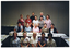 COL photo of a group of people gathered around a long rectangular table. Four people appear to be kneeling on the floor, in front of the table. The remainder of the group are standing behind the table in three rows: the first row of people are sitting on chairs, and the last two rows of people are standing behind them. There are a few other tables around the room. On top of the tables, there are folders, books, and other printed materials. In the background, there is a set of closed bi-fold doors forming a wall at the back.