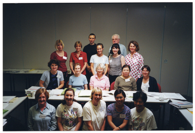 COL photo of a group of people gathered around a long rectangular table. Four people appear to be kneeling on the floor, in front of the table. The remainder of the group are standing behind the table in three rows: the first row of people are sitting on chairs, and the last two rows of people are standing behind them. There are a few other tables around the room. On top of the tables, there are folders, books, and other printed materials. In the background, there is a set of closed bi-fold doors forming a wall at the back.