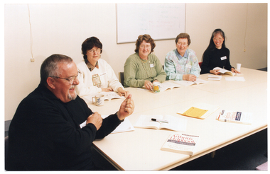 COL photo of a small group of people sitting around a long rectangular table. One man is sitting at the end of the table, while four women are sitting along one side of the table. On the table, there are two closed books depicting the title, "The Arthritis Helpbook", and each person has an open book on the table in front of them. There are also a few mugs and a couple of pieces of paper on the table. Behind the women, there is a whiteboard on the wall.