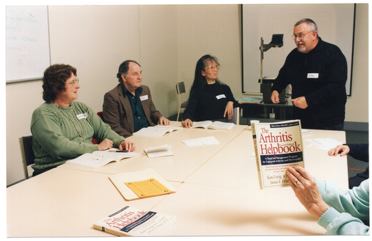 COL photo of a small group of people sitting or standing around four rectangular tables which have been pushed together. One man is standing up at one end of the tables, smiling and gesticulating. The remainder of the group are seated around three sides of the group of tables (two partially obscured). One person (partially obscured) is holding up a book titled, "The Arthritis Helpbook". On the table, there is another book with the same title and some printed paper materials. Three other people each have an open book on the table in front of them. In the background, there is a slide projector and some blue paperwork on a small table, with a projector screen affixed to the wall behind it. On a perpendicular wall, there is a whiteboard also affixed to the wall.