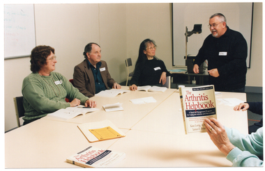 COL photo of a small group of people sitting or standing around four rectangular tables which have been pushed together. One man is standing up at one end of the tables, smiling and gesticulating. The remainder of the group are seated around three sides of the group of tables (two partially obscured). One person (partially obscured) is holding up a book titled, "The Arthritis Helpbook". On the table, there is another book with the same title and some printed paper materials. Three other people each have an open book on the table in front of them. In the background, there is a slide projector and some blue paperwork on a small table, with a projector screen affixed to the wall behind it. On a perpendicular wall, there is a whiteboard also affixed to the wall.