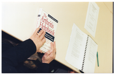 COL photo of a person's hands on top of a book titled, "The Arthritis Helpbook". The book is sitting on top of a table. Next to the book, there is a ring-bound printed training manual, a printed sheet of paper, and a pen.